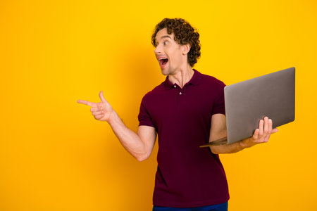 Cheerful man with curly hair showcasing a laptop while pointing energetically against a vibrant yellow backgroundの写真素材