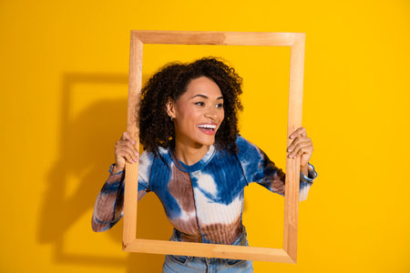 Portrait of a cheerful woman holding a frame on a yellow background, expressing joy and vibrant energyの写真素材