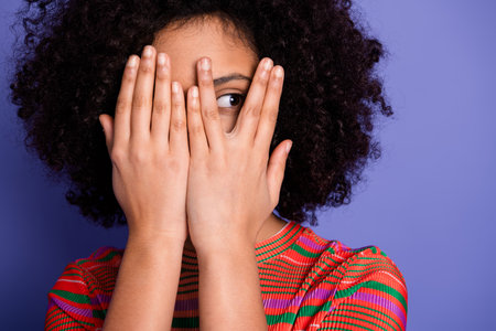 Young girl with curly hair peeking through her fingers in a stylish t-shirt against a vibrant purple...