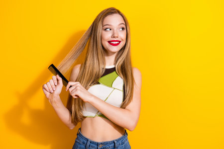 Smiling young woman with long blond hair combing it while standing against a bright yellow...