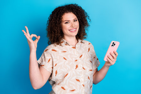 Young woman with curly hair holding smartphone smiling confidently against a blue background while...