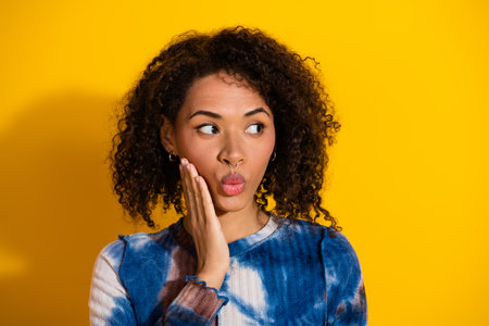 Attractive young woman with curly hair looking surprised against vibrant yellow background wearing a...