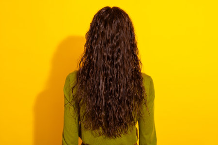 Rear view of a woman with long curly hair in green shirt against a yellow background highlighting modern fashionの写真素材