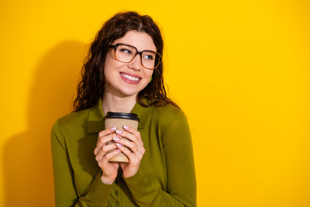 Young cheerful woman wearing glasses holding a coffee cup on a stylish yellow background, radiating...