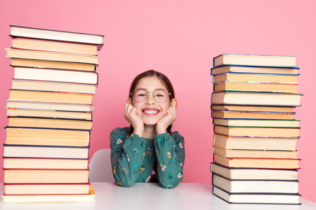 Young girl smiling between stacks of book with a cheerful expression and pink backgroundの写真素材