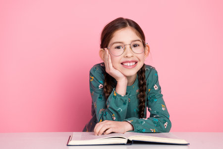 Charming young girl in glasses reading with a cheerful expression against a pink background,...