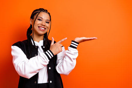 Happy young woman with braided hair pointing and smiling in front of an orange background wearing a...