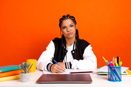 Teenage girl with braids studies at a colorful desk with an orange background, symbolizing education...