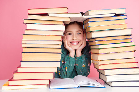 Cheerful young girl surrounded by stacks of colorful book, smiling brightly against a vibrant pink backgroundの写真素材