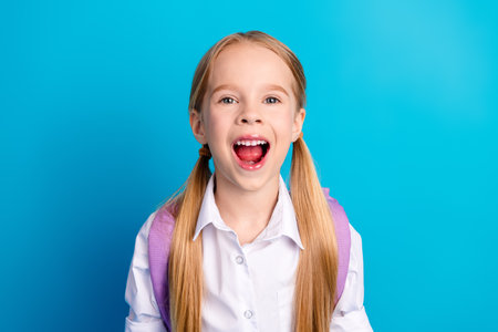 Happy schoolgirl with blonde ponytails wearing a white shirt smiling joyfully on a blue background...
