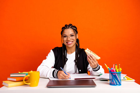 Young student enjoying a snack while studying at a desk with book and supplies in a vibrant orange...