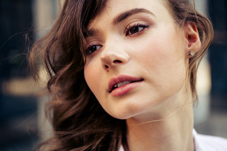 Charming young woman with brunette hair looking into distance during sunny day in urban street environmentの写真素材