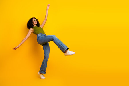 Young stylish woman in green top and jeans on yellow background, showcasing casual fashion, curly hair, and cheerful expressionの写真素材
