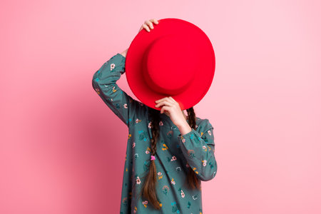 Young girl wearing a green floral dress hiding face with a red hat against pink background expressing playful moodの写真素材