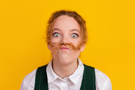 Youthful teen girl making a playful mustache gesture with her hair on a vibrant yellow backgroundの写真素材