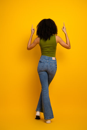 Stylish young woman with curly hair posing on a vibrant yellow background, wearing casual jeans and a green topの写真素材