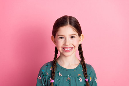 Adorable young girl smiling with braided hairstyle against a pink backdrop, expressing joyful...