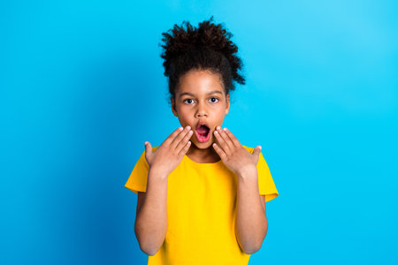 Surprised young girl in a yellow shirt expresses amazement with gesture on blue background, exuding...