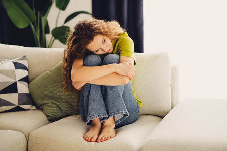 Sad woman with red curly hair sits on a cozy couch indoors, portraying relaxation, comfort, and peaceful thoughtsの写真素材