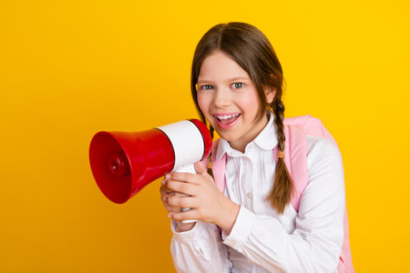 Cheerful schoolgirl holds a red megaphone against a vibrant yellow background, perfect for promotional campaignsの写真素材