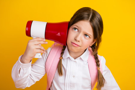 Preteen schoolgirl holding a red megaphone against a bright yellow background showing curious expression in stylish uniformの写真素材