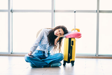Young woman resting at airport terminal with suitcase and travel essentials, dressed casually for a vacation or journey.の写真素材