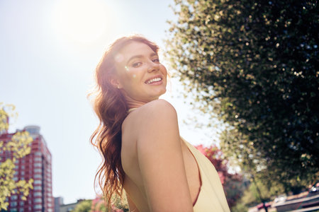 Young woman enjoying a sunny day outdoors in a stylish dress, smiling happily in an urban park settingの写真素材