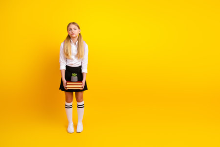 Young schoolgirl holding books and plant against a yellow background, feeling contemplativeの写真素材