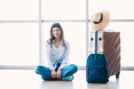 Charming woman at airport sitting with luggage, displaying a cheerful attitude, signifying travel, adventure, or vacationの写真素材