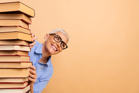 Elderly woman peeking around a stack of book against a beige background, wearing glasses and a striped shirt, smiling joyfully.の写真素材