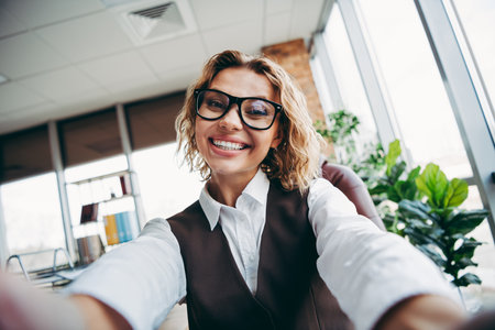 Smiling confident businesswoman in elegant formal attire taking a selfie in a modern office with natural indoor lightの写真素材