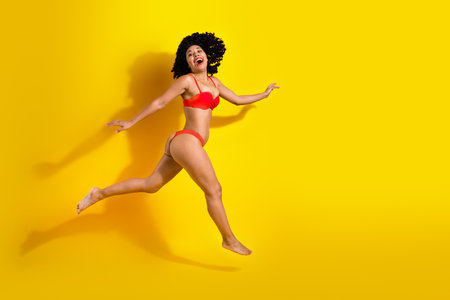 Young woman in vibrant red swimsuit jumping joyfully against yellow background, expressing happiness and summer vibesの写真素材