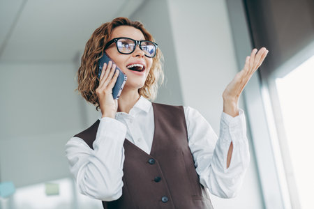 Confident businesswoman engaging in a phone conversation inside an office, showcasing professionalism and modern workplace attireの写真素材