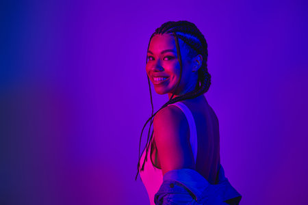 Lovely woman with braided hair in neon lighting, wearing a denim jacket and smiling confidently...