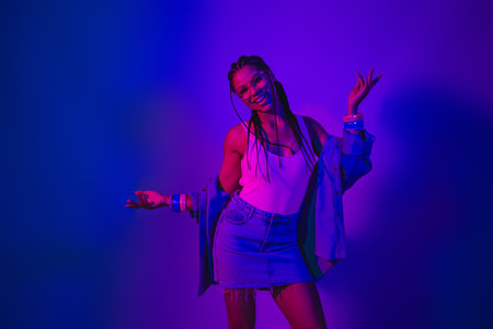 Stylish young woman in neon lighting shows positive gestures wearing modern casual denim fashion for...