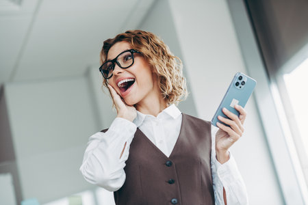 Confident businesswoman celebrating success with smartphone in modern office setting, showcasing professional attire and enthusiasmの写真素材