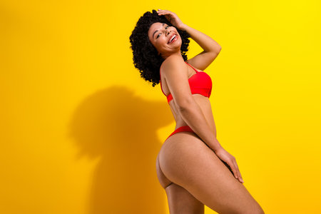 Confident young woman in red swimwear posing energetically against a vibrant yellow background, expressing happinessの写真素材