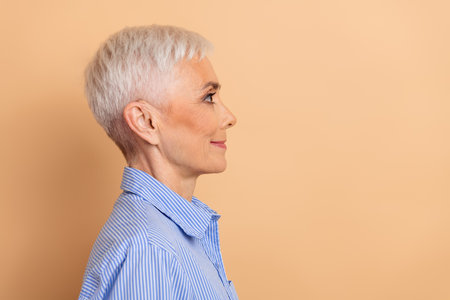 Profile view of confident mature woman with short white hair wearing blue striped shirt on beige...