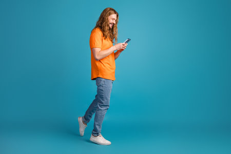 Young man walking casually while using a smartphone against a vibrant blue background showcasing leisure and technologyの写真素材