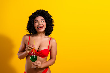 Fashionable young woman in red bikini with curly hair holding a drink and smiling against a vibrant yellow backgroundの写真素材