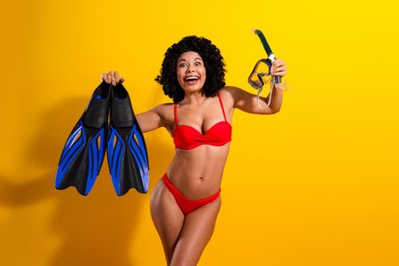 Vibrant joyful woman in red bikini holding snorkeling gear against yellow background, embracing summer adventures and beach funの写真素材