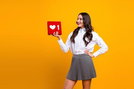 Charming stylish female holding red like symbol on yellow background showing hand gesture wearing...