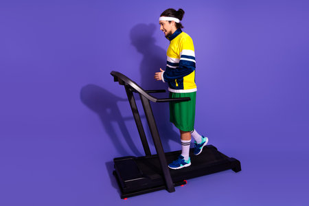 Active man in bright retro sportswear walking on a treadmill against a violet background showcasing...