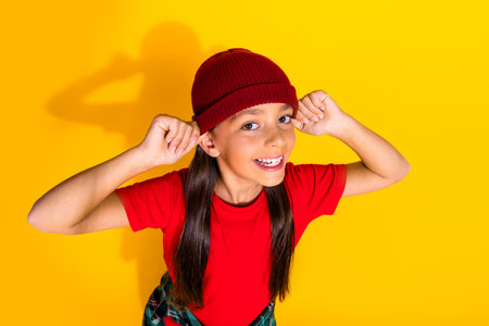 Happy young girl posing playfully in a knitted beanie against a vibrant yellow background, expressing happinessの写真素材