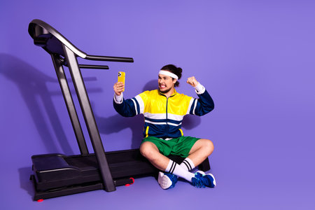 Young athlete in retro sportswear posing with smartphone near treadmill on vibrant purple background
