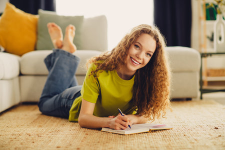 Cute woman with curly hair writing in a notebook while lying on a rug in a cozy living room with...