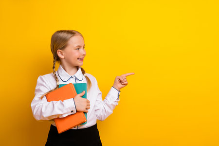 Cheerful schoolgirl holding book against a yellow background pointing to the side promoting education and learningの写真素材