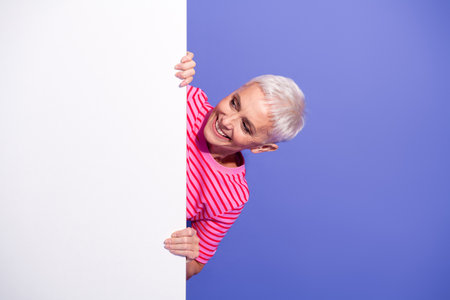 Happy elderly woman in striped pink shirt peeking around blank space with cheerful expression against purple backgroundの写真素材