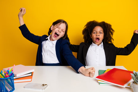 Two cheerful schoolgirls dressed in formal attire stretching and smiling in a colorful classroom settingの写真素材