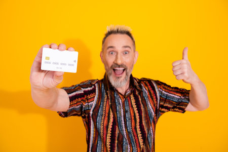 Mature man showing credit card and giving thumbs up against vibrant yellow background, representing...